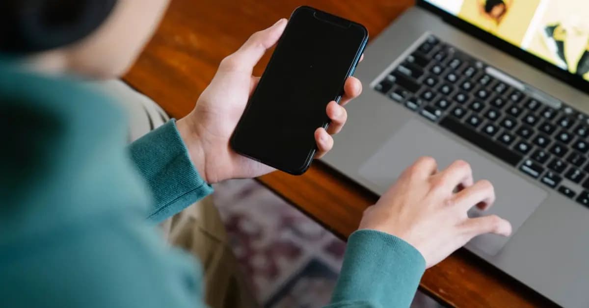 Person using a laptop while holding a smartphone in a home office